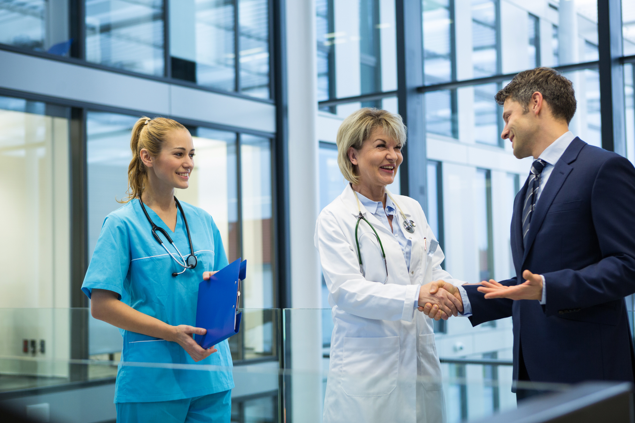 Female doctor providing physician coverage is shaking hands with businessman as healthcare worker in scrubs looks on. 
